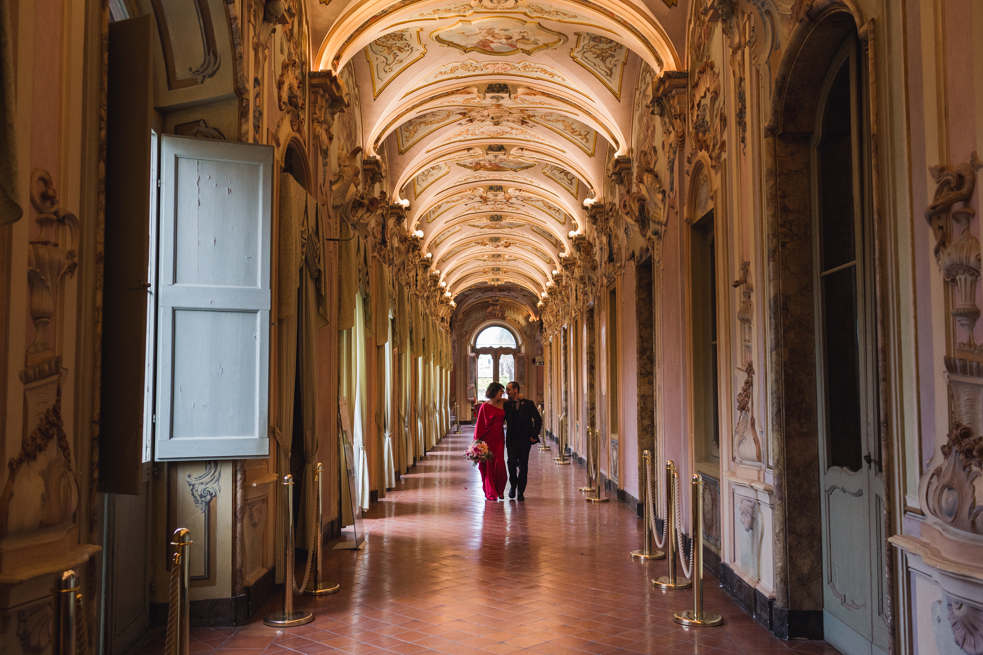 Galleria degli stucchi con due sposi che stanno camminando abbracciati. La sposa è vestita di rosso e tiene un mazzo di fiori in tinta con l'abito.